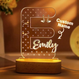 May include: A clear acrylic earring holder shaped like the letter "E", illuminated with warm light. The base is made of wood. The name "Emily" is written in a cursive font. The holder has several holes for earrings and displays a pair of butterfly earrings.