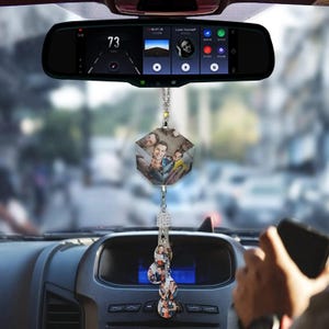 Puede incluir: Un espejo retrovisor de coche con un adorno fotográfico colgante. El adorno tiene forma de múltiples caras con fotos familiares. Debajo, pequeños colgantes con fotos. Se ve el salpicadero del coche y una mano sosteniendo un teléfono.