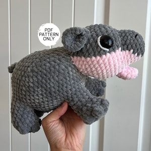 Op de afbeelding: Een handgemaakte gehaakte nijlpaard knuffel in grijs en roze. Het nijlpaard heeft een grijze body, een roze snuit en mond, en grote zwart-witte ogen. De tekst "PDF PATTERN ONLY" staat in een witte cirkel.
