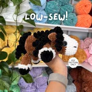 May include: A handmade plush horse with brown, black, and white details. The text "LOW-SEW!" is above the horse, and "PDF PATTERN ONLY" is below. The horse is held in front of a shelf with colorful yarn.