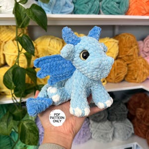 Könnte beinhalten: Ein hellblauer gehäkelter Drache mit großen, runden, goldenen Augen, blauen Flügeln und weißen Akzenten an den Füßen. Der Drache ist aus weichem, plüschigem Garn gefertigt. Das Bild enthält den Text "PDF PATTERN ONLY".