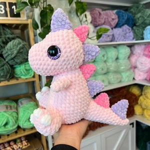 Puede incluir: Un juguete de dinosaurio de crochet rosa y morado con un ojo morado brillante. El dinosaurio est&aacute; sentado en una mano.