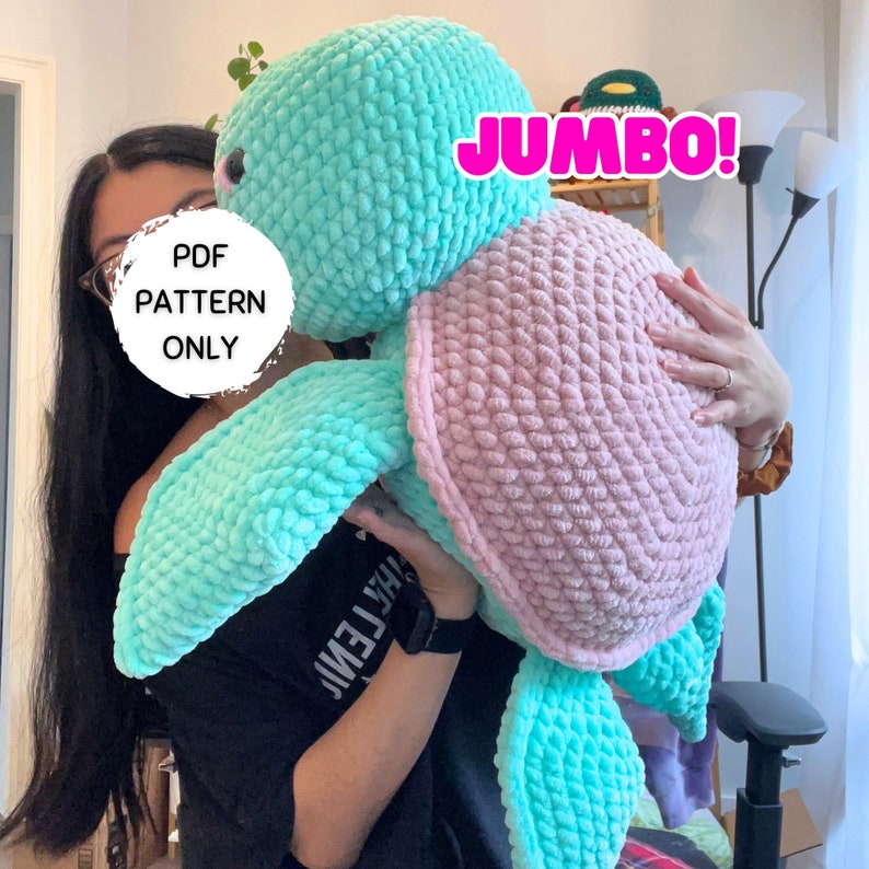 May include: A large crocheted turtle amigurumi toy in shades of pink and turquoise. The toy is being held by a person. The text "JUMBO!" is visible in the image. The text "PDF PATTERN ONLY" is also visible in the image.