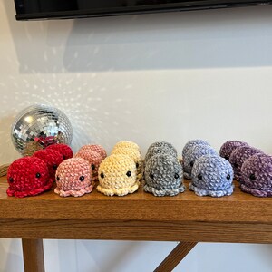 Op de afbeelding: Een rij van 10 gehaakte octopus knuffels in een regenboog van kleuren, van rood tot paars, op een houten tafel.