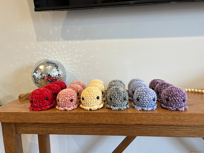 Op de afbeelding: Een rij van 10 gehaakte octopus knuffels in een regenboog van kleuren, van rood tot paars, op een houten tafel.