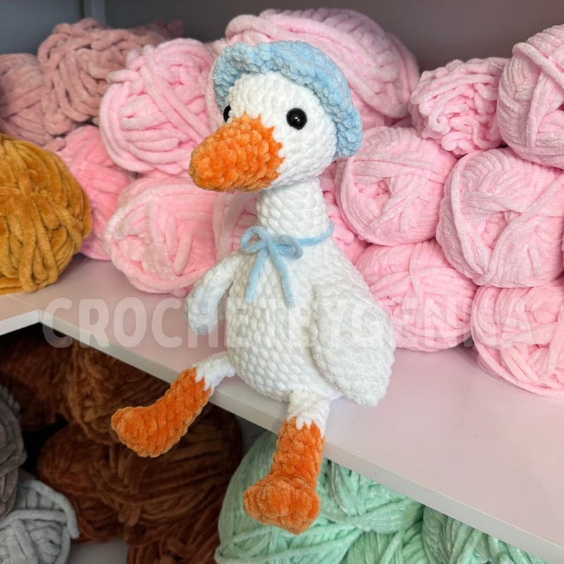 May include: A white crocheted plush goose with an orange beak and feet, wearing a light blue crocheted bonnet and a matching light blue ribbon tied around its neck. The goose has black plastic eyes and a soft, textured yarn construction.