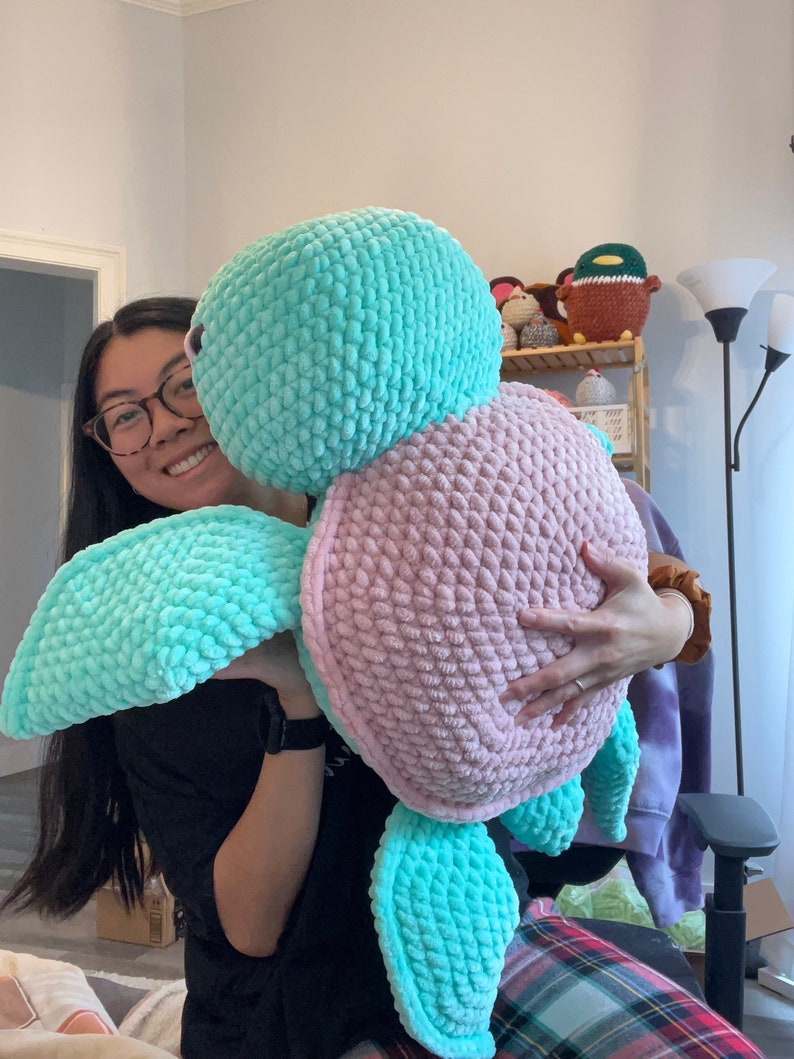 May include: A person holding a large crocheted stuffed turtle. The turtle is pink and blue with a textured surface.