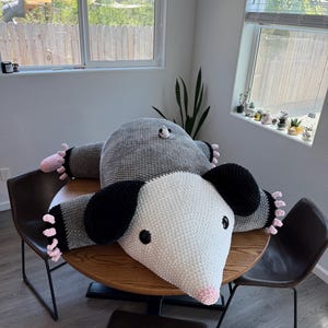 May include: Two large, handmade crochet possum pillows in gray and white, with black ears and pink accents. The pillows are lying on a round wooden table, with black chairs surrounding it. The possums have black eyes and pink noses.
