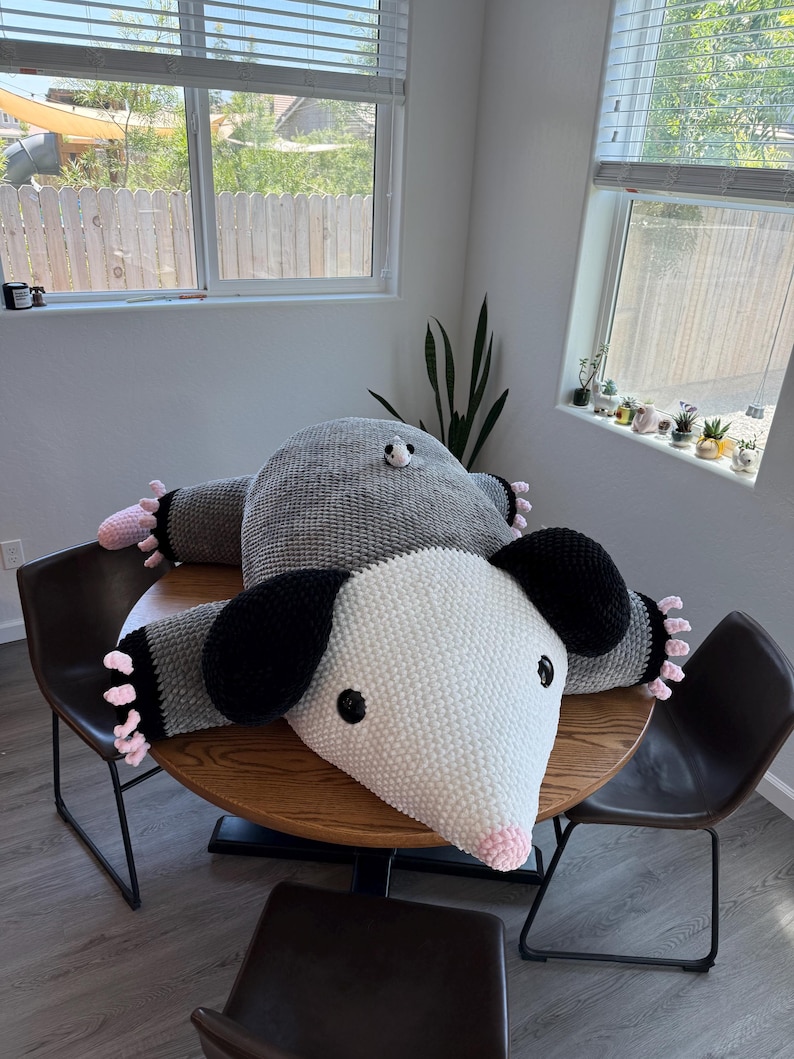 May include: Two large, handmade crochet possum pillows in gray and white, with black ears and pink accents. The pillows are lying on a round wooden table, with black chairs surrounding it. The possums have black eyes and pink noses.