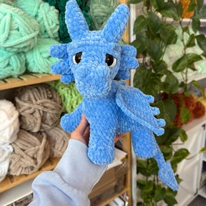 K&ouml;nnte beinhalten: Ein hellblauer geh&auml;kelter Drache mit gro&szlig;en Augen, Fl&uuml;geln und einem langen Schwanz. Das Pl&uuml;schtier ist aus weichem, strukturiertem Garn gefertigt. Der Drache wird vor Regalen mit verschiedenen Garnfarben pr&auml;sentiert.