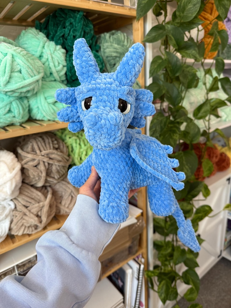 K&ouml;nnte beinhalten: Ein hellblauer geh&auml;kelter Drache mit gro&szlig;en Augen, Fl&uuml;geln und einem langen Schwanz. Das Pl&uuml;schtier ist aus weichem, strukturiertem Garn gefertigt. Der Drache wird vor Regalen mit verschiedenen Garnfarben pr&auml;sentiert.