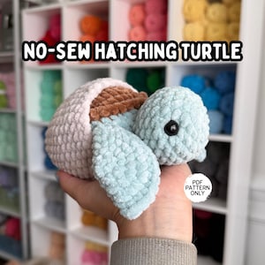 Puede incluir: Una tortuga de ganchillo en tonos azul claro, marrón y blanco. La tortuga tiene un aspecto texturizado y afelpado, con una cabeza redonda y apéndices en forma de aleta. El texto "NO-SEW HATCHING TURTLE" se muestra encima de la tortuga.