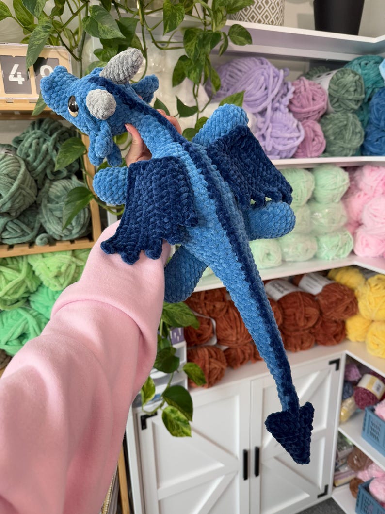 May include: A blue crocheted dragon toy with a long tail and wings. The dragon is being held by a hand wearing a pink sweater. The dragon is in front of a shelf with yarn balls in various colors.