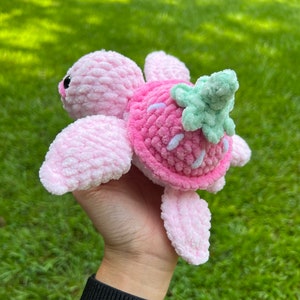 May include: A pink crocheted turtle with a strawberry shell and green leaves. The turtle is held in a hand with a black wristband.