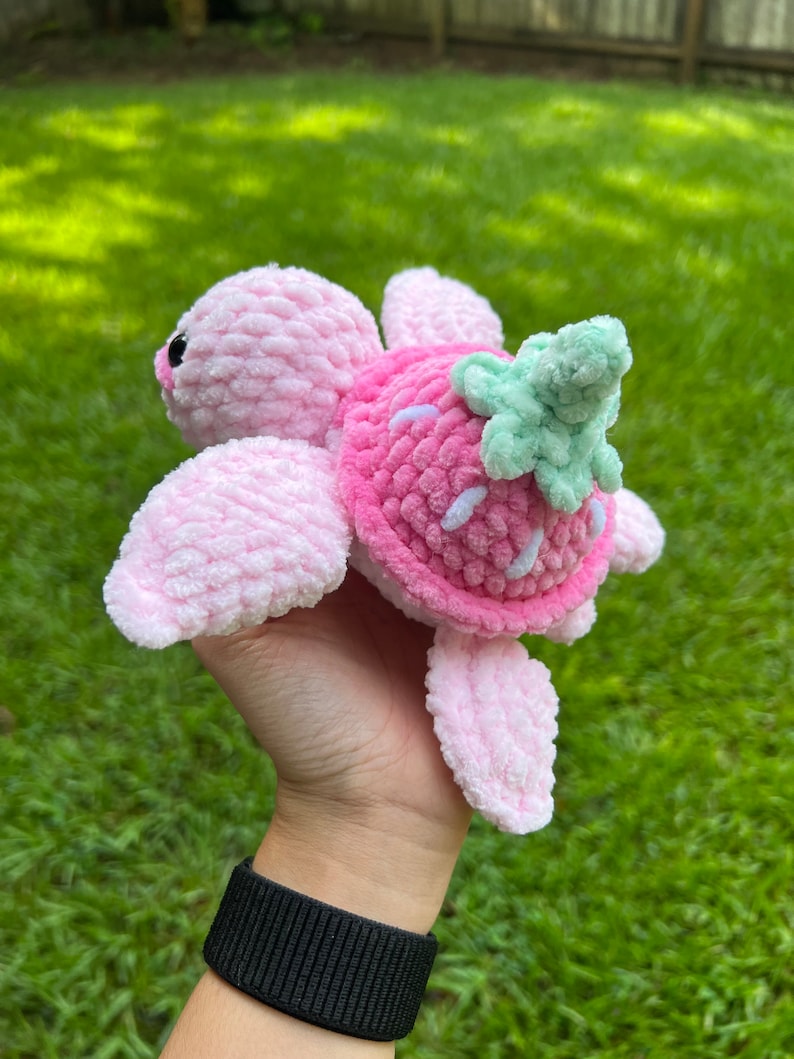 May include: A pink crocheted turtle with a strawberry shell and green leaves. The turtle is held in a hand with a black wristband.