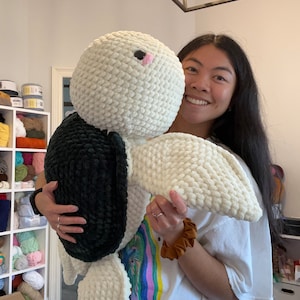 May include: A person holding a large white and black crocheted stuffed animal. The animal has a pink nose and black eyes. The person is wearing a white t-shirt with a colorful design.