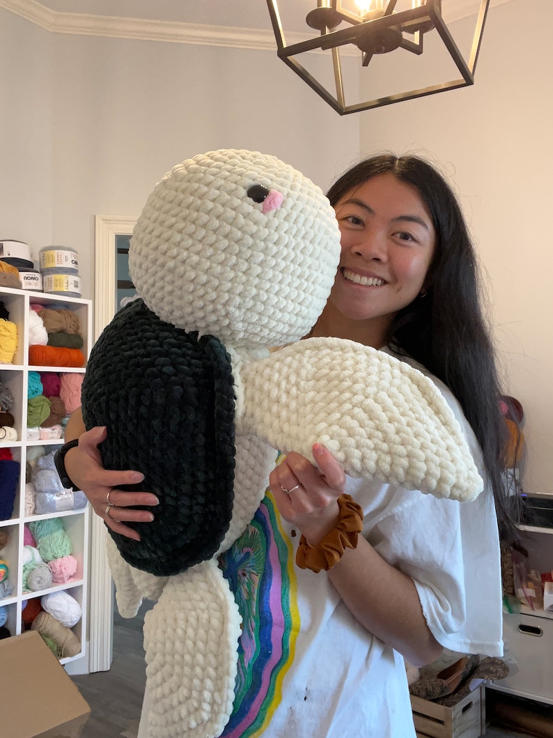 May include: A person holding a large white and black crocheted stuffed animal. The animal has a pink nose and black eyes. The person is wearing a white t-shirt with a colorful design.
