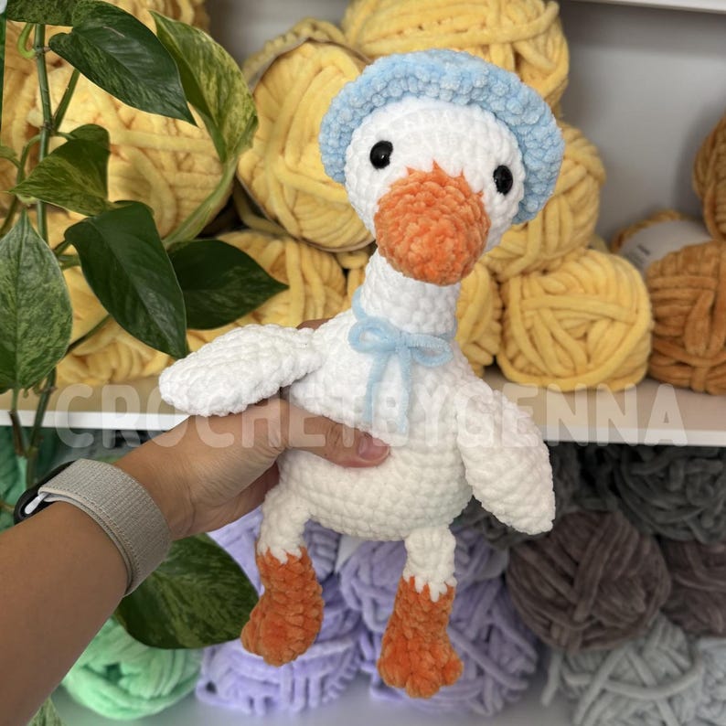 May include: A white plush crochet goose toy with an orange beak and feet, wearing a light blue textured bonnet and a matching light blue ribbon tied around its neck. The toy features black safety eyes and a soft, fuzzy yarn construction.