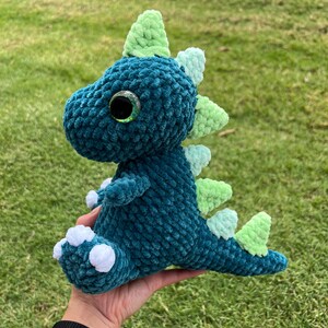 Puede incluir: Un juguete de dinosaurio de crochet verde azulado con una espalda con p&uacute;as verde y blanca y pies blancos. El dinosaurio tiene un ojo verde grande con una pupila negra.