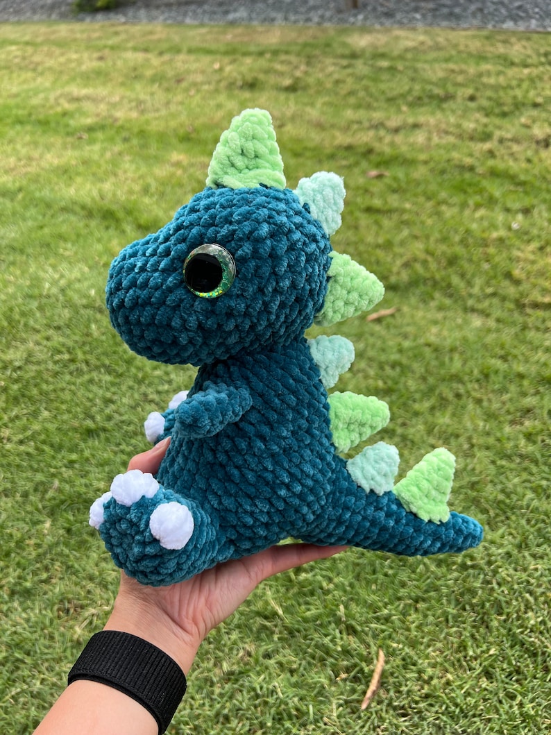 Puede incluir: Un juguete de dinosaurio de crochet verde azulado con una espalda con p&uacute;as verde y blanca y pies blancos. El dinosaurio tiene un ojo verde grande con una pupila negra.