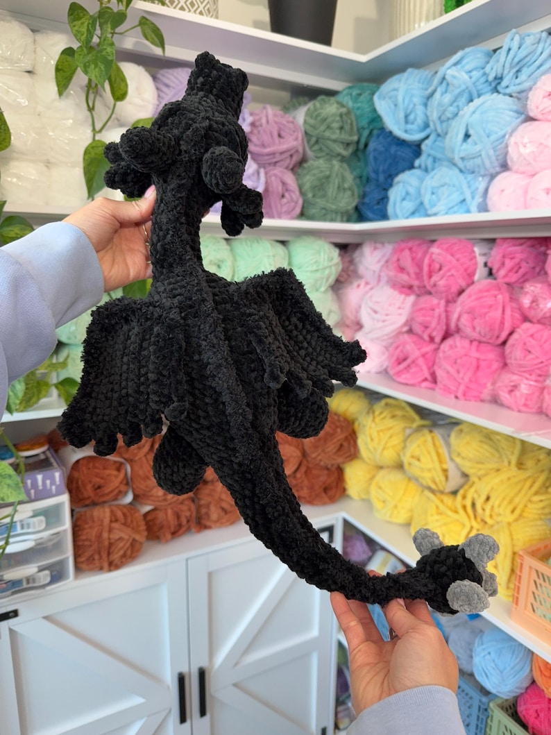 May include: A black crocheted dragon toy with gray claws. The dragon is posed with its wings spread out and its tail curled around its body.