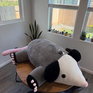 May include: A large, handmade crochet opossum plush toy in shades of gray, white, black, and pink. The plush is lying on a wooden table. A small crochet mouse is on the opossum's back. The toy has black ears and pink feet.