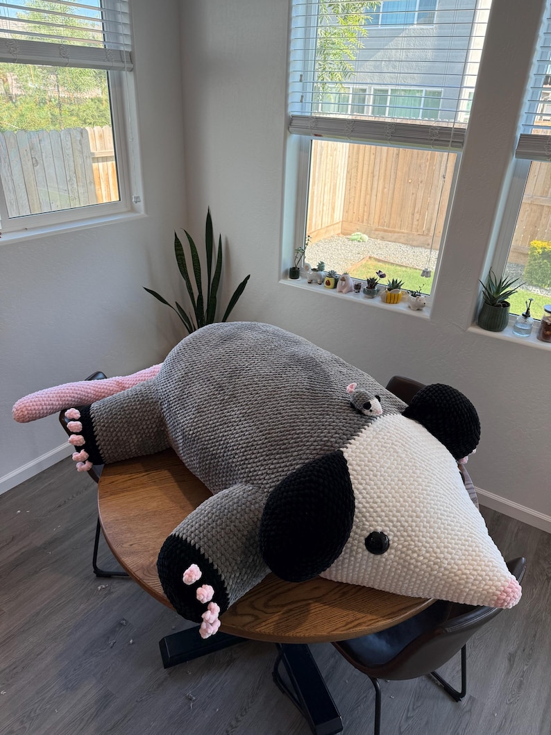 May include: A large, handmade crochet opossum plush toy in shades of gray, white, black, and pink. The plush is lying on a wooden table. A small crochet mouse is on the opossum's back. The toy has black ears and pink feet.