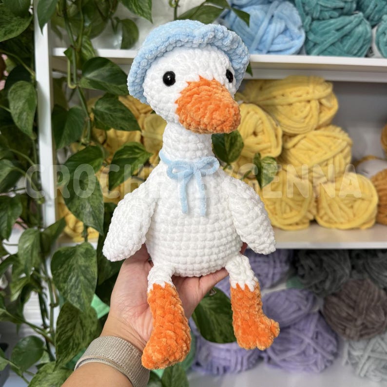 May include: A handmade crochet plush goose featuring a white body, bright orange feet and beak, and a light blue textured bonnet. The goose has black bead eyes and a small light blue ribbon tied around its neck, crafted from soft yarn.