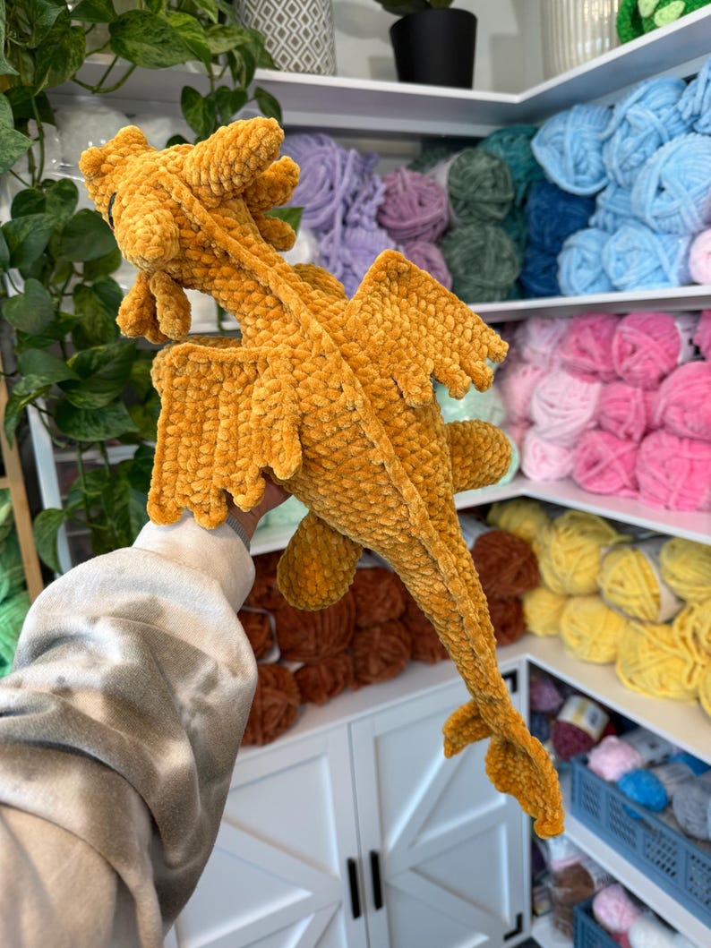 May include: A crocheted plush dragon toy in a mustard yellow color. The dragon has wings and a long tail. The toy is being held by a hand in front of a shelf of yarn.