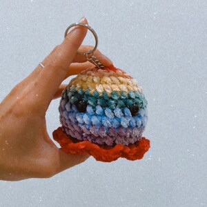 Op de afbeelding: Een regenboogkleurige gehaakte octopus sleutelhanger met zwarte ogen en een rode tentakel.
