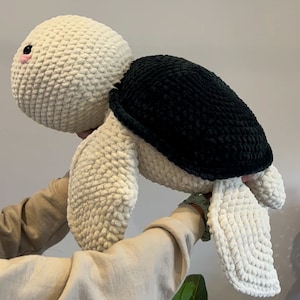 May include: A crocheted stuffed turtle toy with a white body and black shell. The turtle has a pink nose and is being held by a person's hands.