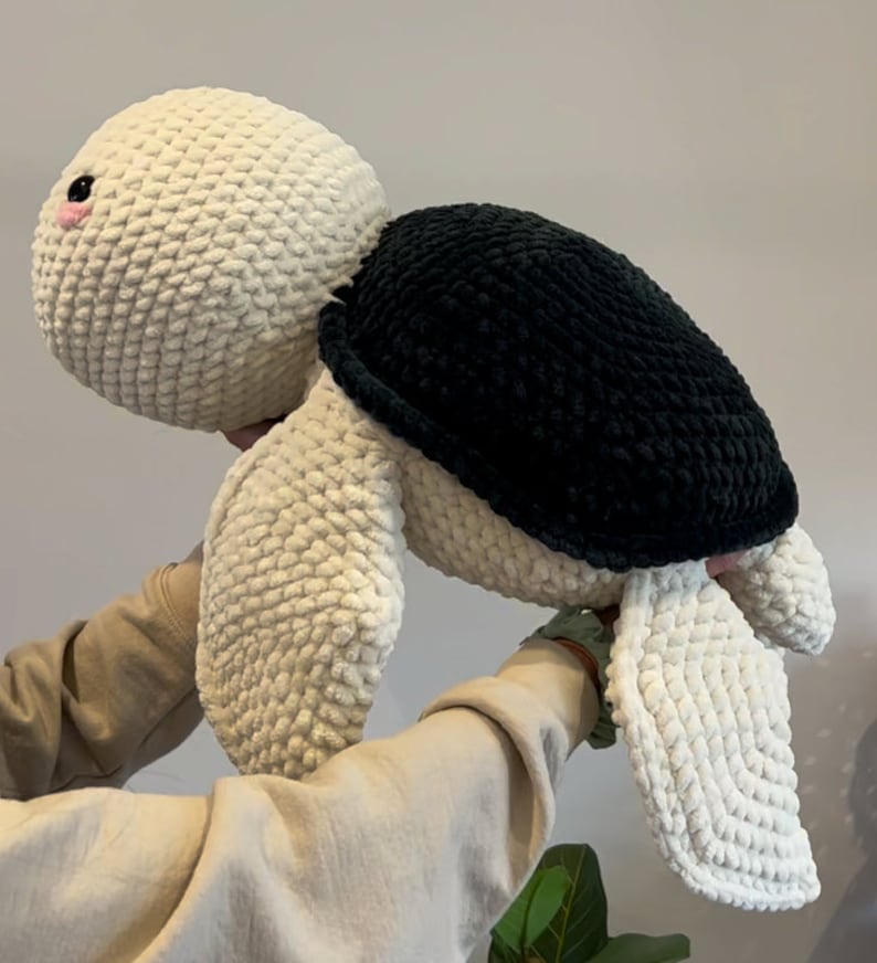 May include: A crocheted stuffed turtle toy with a white body and black shell. The turtle has a pink nose and is being held by a person's hands.