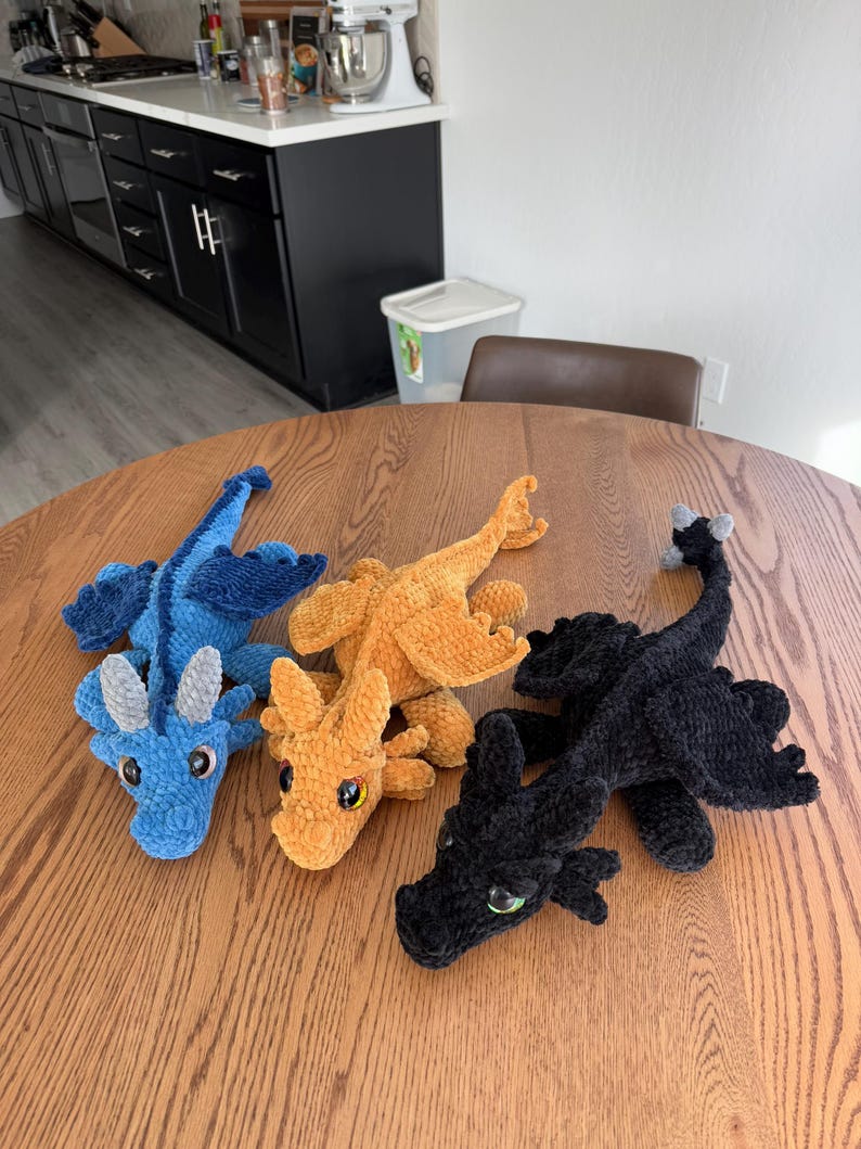 May include: Three crocheted plush dragons on a wooden table. The dragons are blue, orange, and black.
