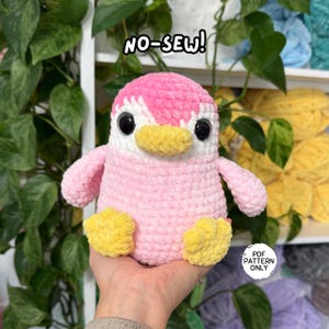 Op de afbeelding: Een gehaakte pluche pinguïn met een zachtroze lichaam, een gele snavel en gele voeten. De pinguïn heeft grote zwarte veiligheidsogen en een witte vlek op het gezicht. De tekst NO-SEW! is bovenaan zichtbaar en een label vermeldt PDF PATTERN ONLY.