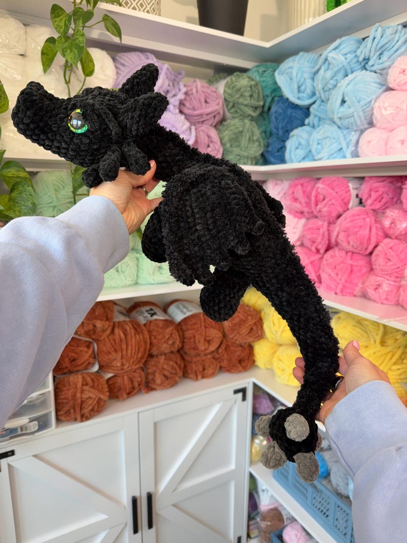May include: A black crocheted dragon plush toy with green eyes. The dragon is being held up by two hands.