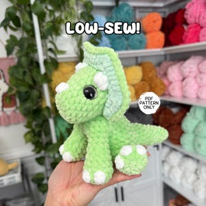 Puede incluir: Un dinosaurio Triceratops de ganchillo verde claro hecho a mano con detalles blancos. El peluche tiene ojos negros grandes y está etiquetado "LOW-SEW!" en texto blanco. El fondo incluye estantes llenos de hilo colorido.