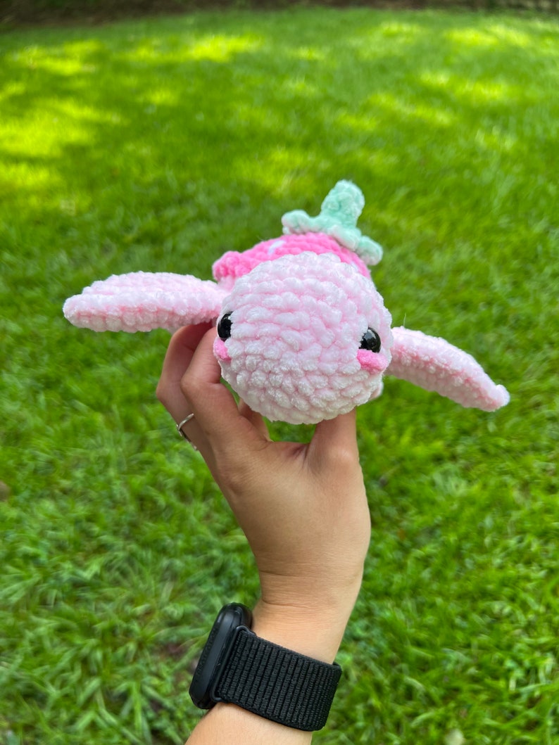 May include: A pink crocheted turtle with a green crocheted shell. The turtle is being held in a hand with a black watch on the wrist.