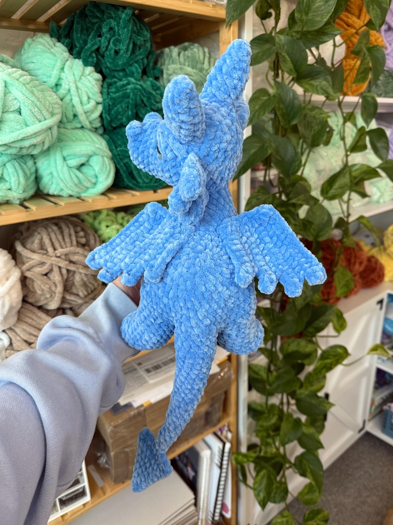 K&ouml;nnte beinhalten: Ein hellblaues, pl&uuml;schiges Drachenspielzeug mit detaillierten Fl&uuml;geln, einem langen Schwanz und einem strukturierten K&ouml;rper. Der handgefertigte Drache wird vor einem Hintergrund aus Garnkn&auml;ueln in verschiedenen Farben, darunter Gr&uuml;n und Beige, pr&auml;sentiert. Der Drache ist etwa 25 cm gro&szlig;.