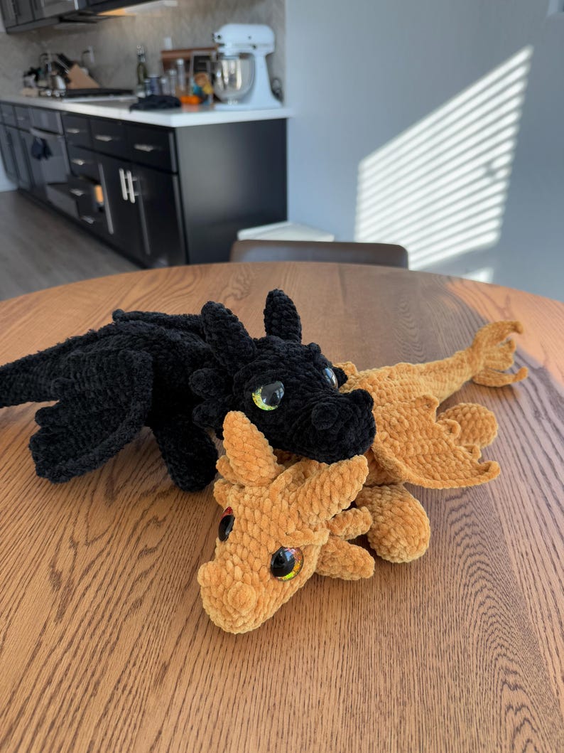 May include: Two crocheted dragon plushies, one black and one brown, with large, colorful eyes. The dragons are lying on a wooden table.