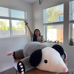 May include: A large, handmade crochet opossum plush toy in shades of gray, white, black, and pink. The plush is lying on a round wooden table. The opossum has black ears, pink paws, and a white face.
