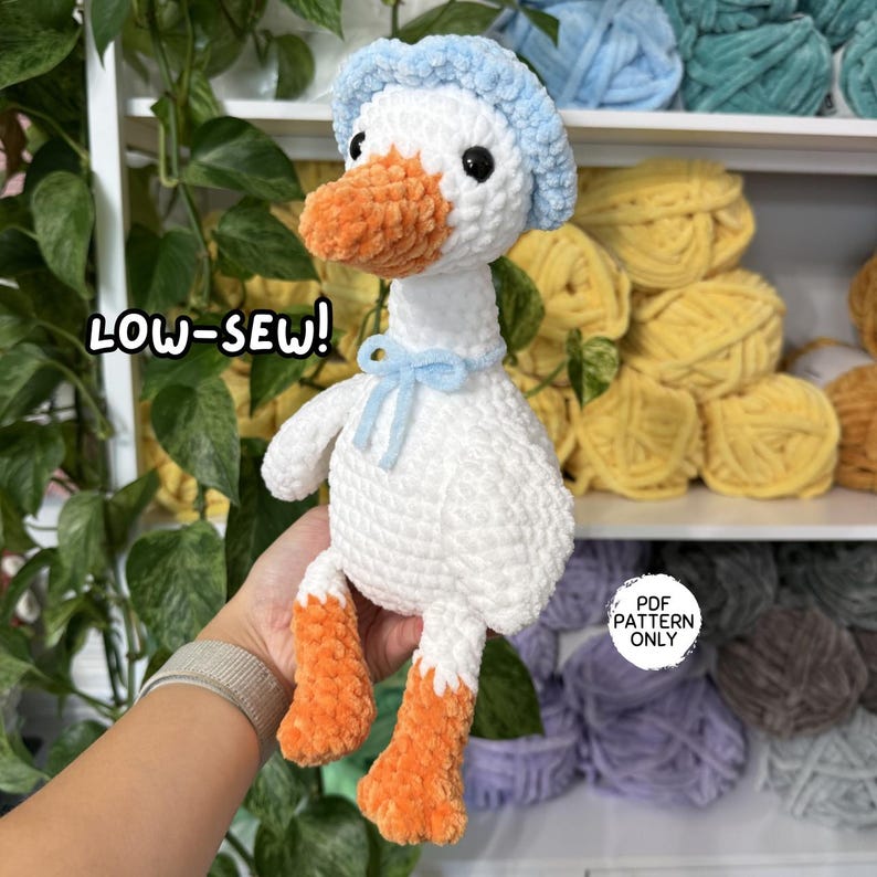 May include: A white crochet goose plushie with an orange beak and feet, wearing a light blue bonnet and a matching blue bow around its neck. The image includes the text LOW-SEW! and PDF PATTERN ONLY. The plushie is made of soft yarn.