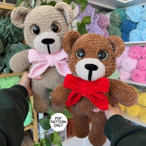 May include: Two crocheted teddy bears, one brown and one beige, with large eyes and a bow. The brown bear has a red bow and the beige bear has a pink bow. The bears are sitting on a table with yarn balls in the background.  PDF PATTERN ONLY.