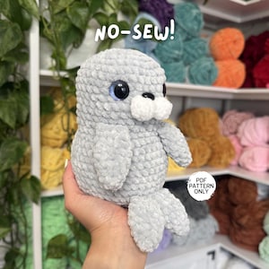 May include: A gray crocheted seal plushie with blue eyes, white accents, and flipper-like appendages. The text "NO-SEW!" is above the seal. The words "PDF PATTERN ONLY" are also visible.