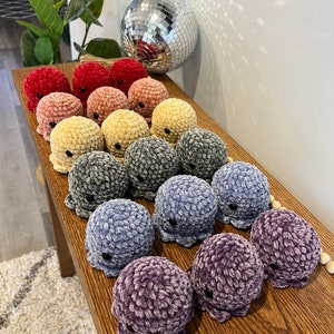 Op de afbeelding: Een verzameling gehaakte octopus knuffels in verschillende kleuren, waaronder rood, roze, geel, grijs, blauw en paars. De knuffels zijn in een rij op een houten oppervlak gerangschikt.