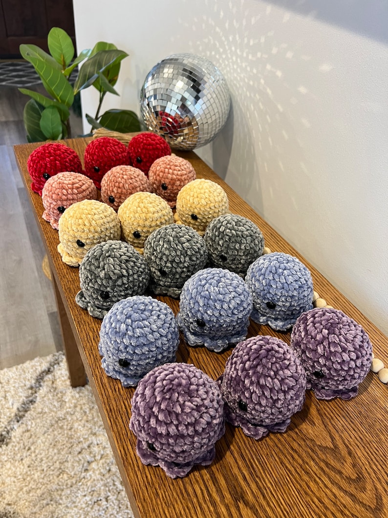 Op de afbeelding: Een verzameling gehaakte octopus knuffels in verschillende kleuren, waaronder rood, roze, geel, grijs, blauw en paars. De knuffels zijn in een rij op een houten oppervlak gerangschikt.