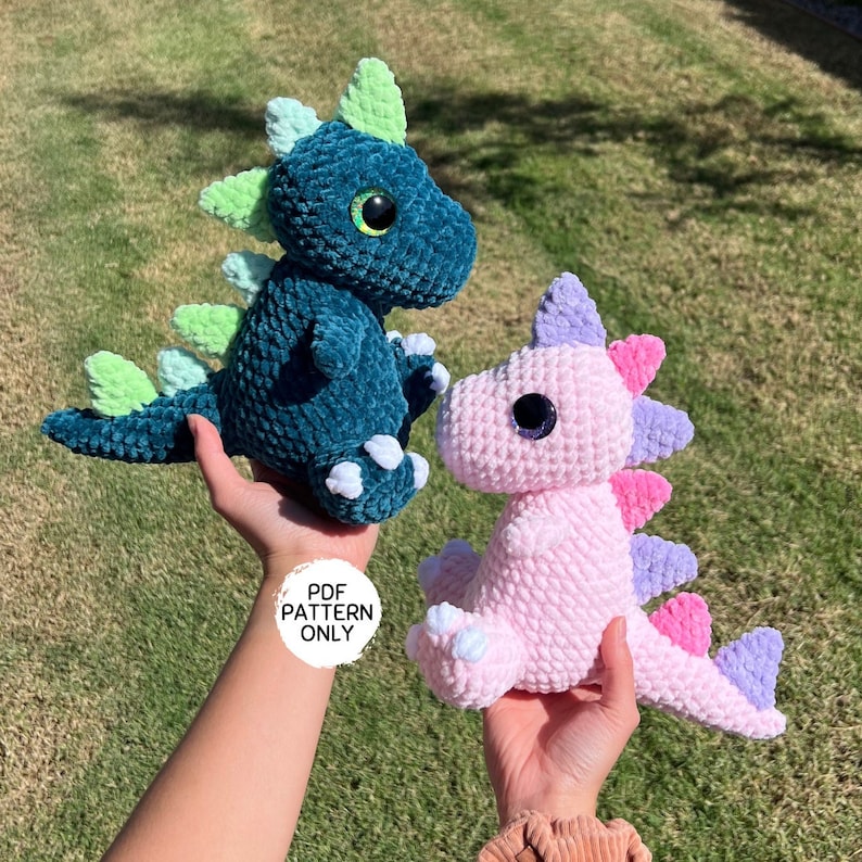 Puede incluir: Dos peluches de dinosaurio de crochet, uno verde azulado con p&uacute;as verde claro y garras blancas, y otro rosa con p&uacute;as moradas y rosas y garras blancas. Los peluches est&aacute;n siendo sostenidos en las manos de una persona. "PDF PATTERN ONLY" est&aacute; escrito en un c&iacute;rculo blanco.
