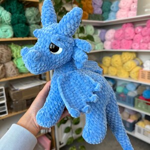 K&ouml;nnte beinhalten: Ein hellblaues, pl&uuml;schiges Drachenspielzeug mit gro&szlig;en, schwarzen Augen, Fl&uuml;geln und einem langen Schwanz. Der Drache besteht aus einem weichen, strukturierten Material. Regale im Hintergrund sind mit farbenfrohem Garn gef&uuml;llt.