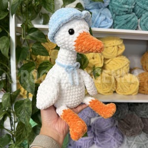 May include: A white crocheted goose plush toy featuring a light blue hat, an orange beak, and orange feet. The goose has a light blue ribbon tied around its neck. The plush is crafted from soft, textured yarn with visible stitching throughout.