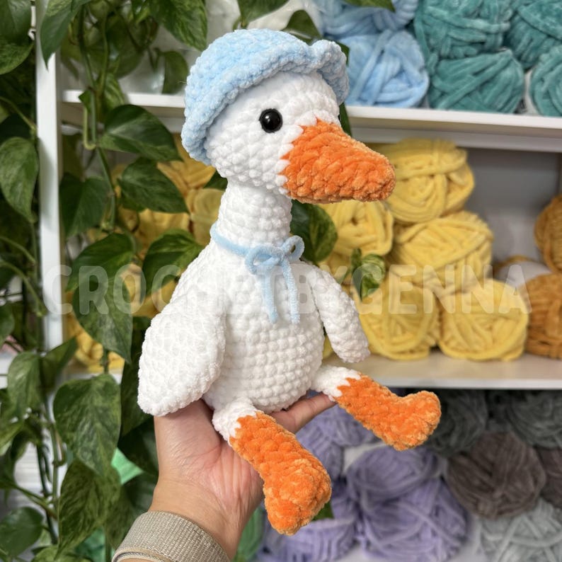 May include: A white crocheted goose plush toy featuring a light blue hat, an orange beak, and orange feet. The goose has a light blue ribbon tied around its neck. The plush is crafted from soft, textured yarn with visible stitching throughout.