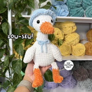 May include: A white crocheted plush goose with an orange beak and feet, wearing a light blue bonnet and a matching blue ribbon around its neck. The text LOW-SEW! is visible on the left, and a circular label reads PDF PATTERN ONLY.